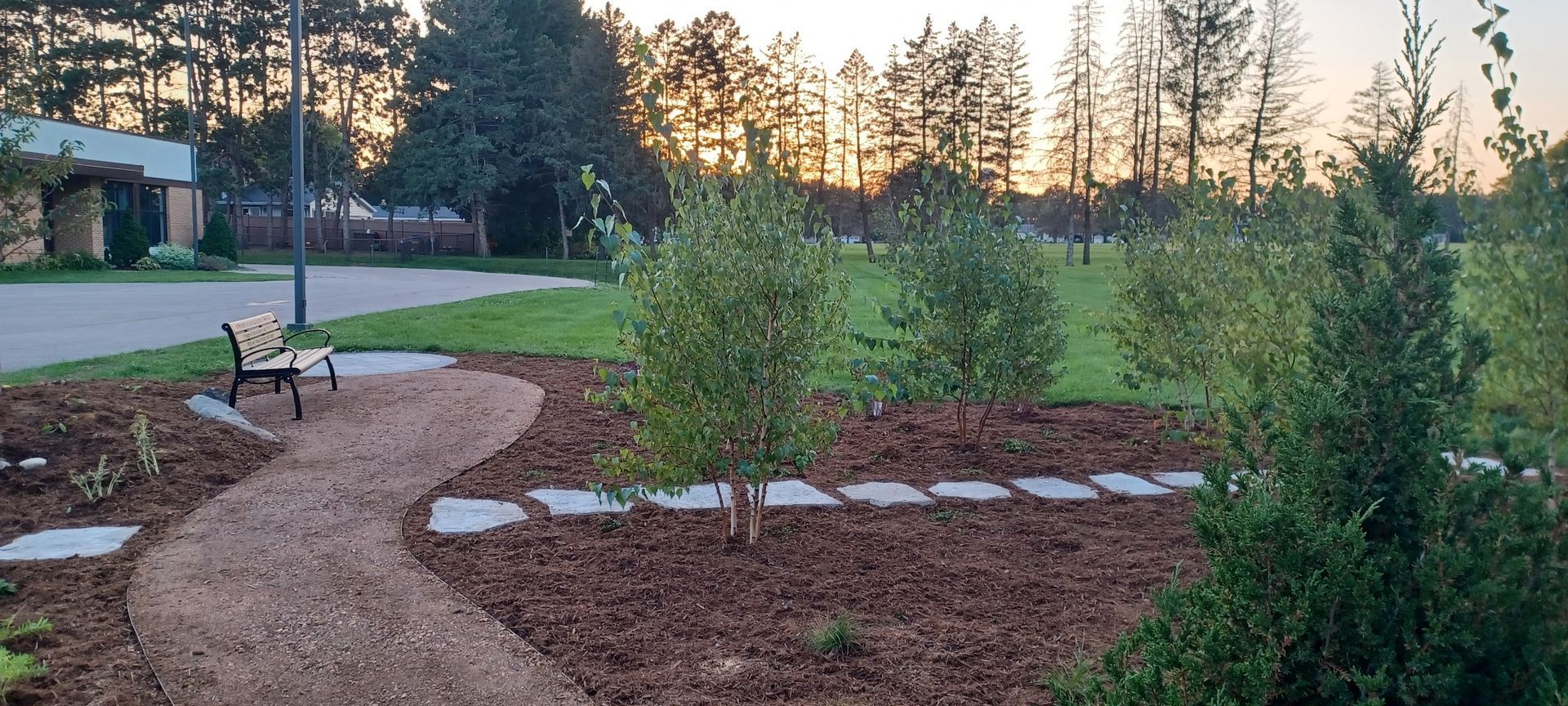 Curved gravel path through a landscaped yard with stepping stones, mulch beds, and young trees at sunset