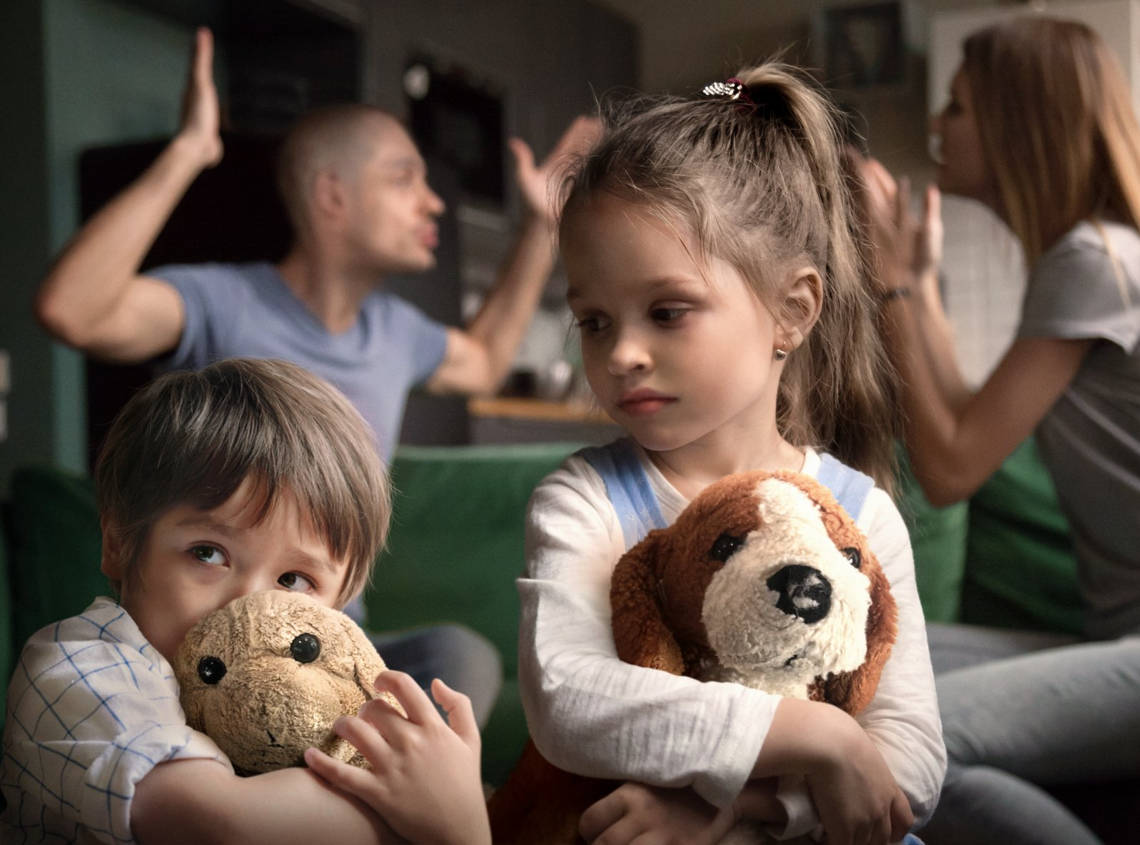 A little girl is holding a stuffed dog while her parents argue in the background.