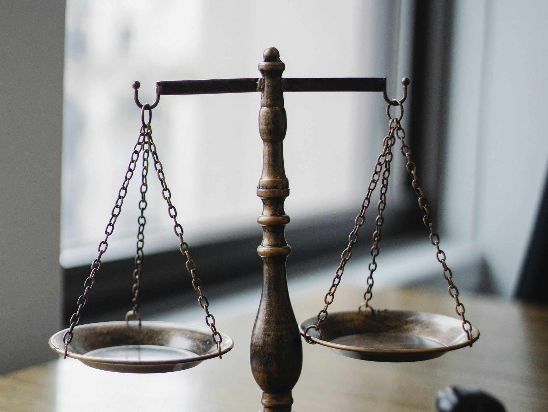 A scale of justice is sitting on a wooden table in front of a window.