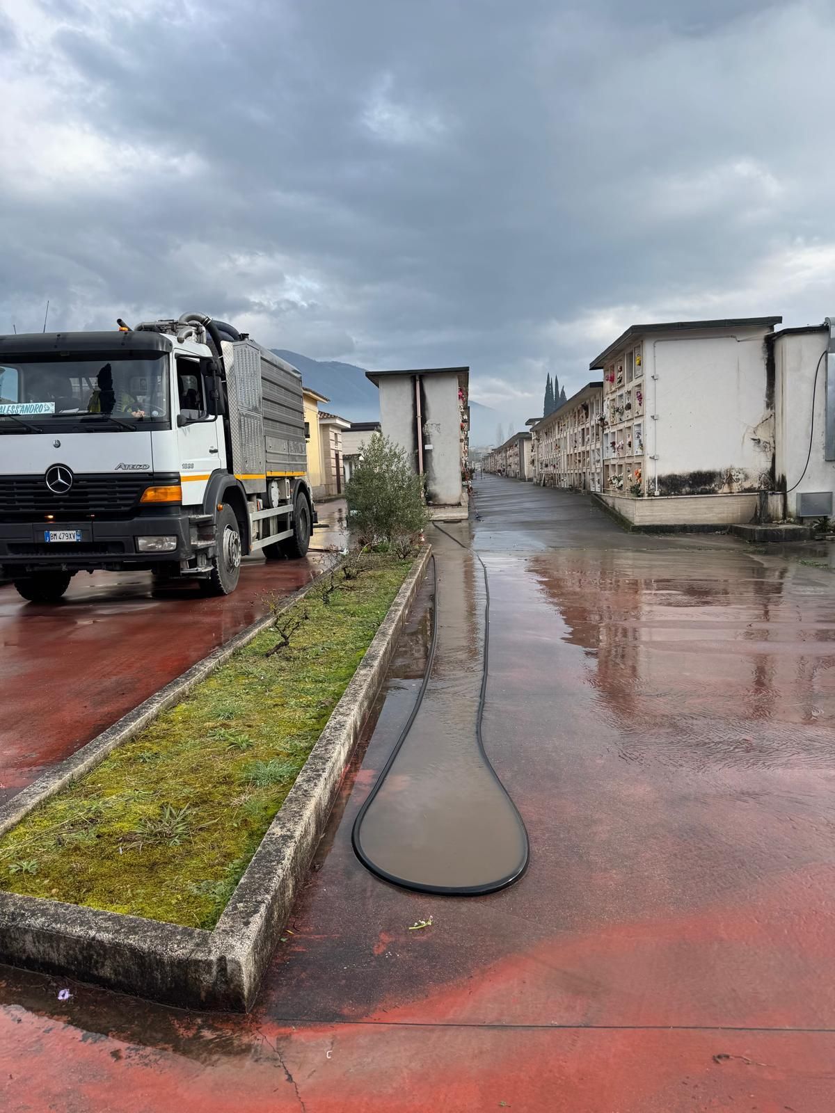 Un camion su una strada bagnata tra edifici bianchi adibiti a magazzino, sotto un cielo nuvoloso.