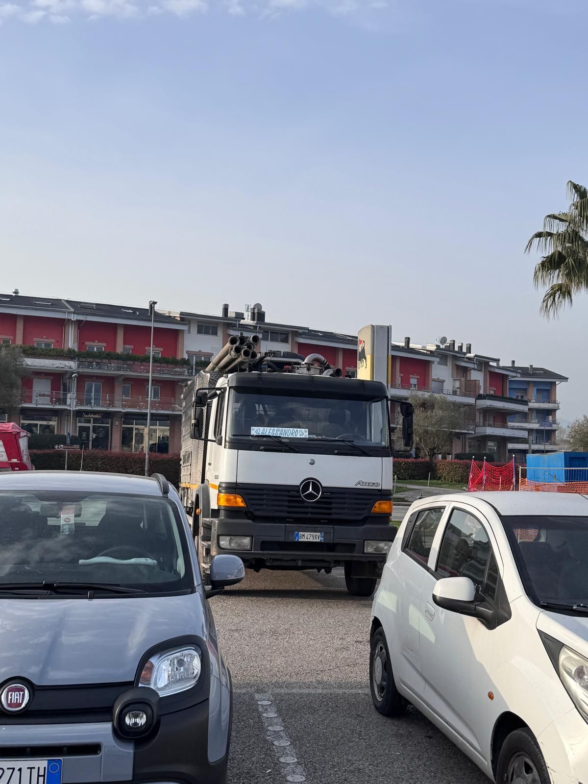 Un grosso camion su una strada tra auto parcheggiate, con edifici e un cielo limpido sullo sfondo.