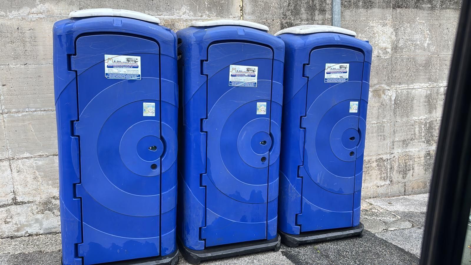 Tre bagni portatili blu allineati contro un muro di cemento all'aperto