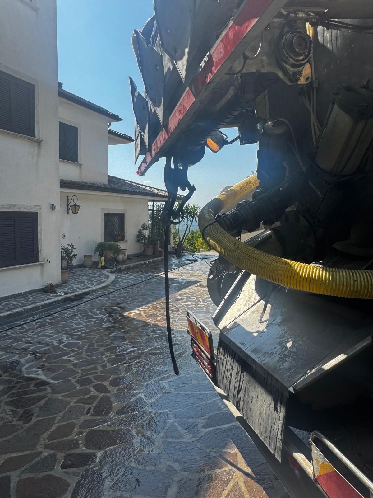 Inquadratura ravvicinata di un grande macchinario nero su una strada acciottolata illuminata dal sole, accanto a edifici bianchi.