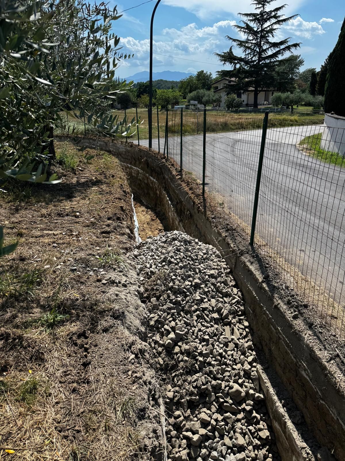 Fossa di drenaggio in ghiaia a lato di una strada con una recinzione metallica, alberi e montagne sullo sfondo.