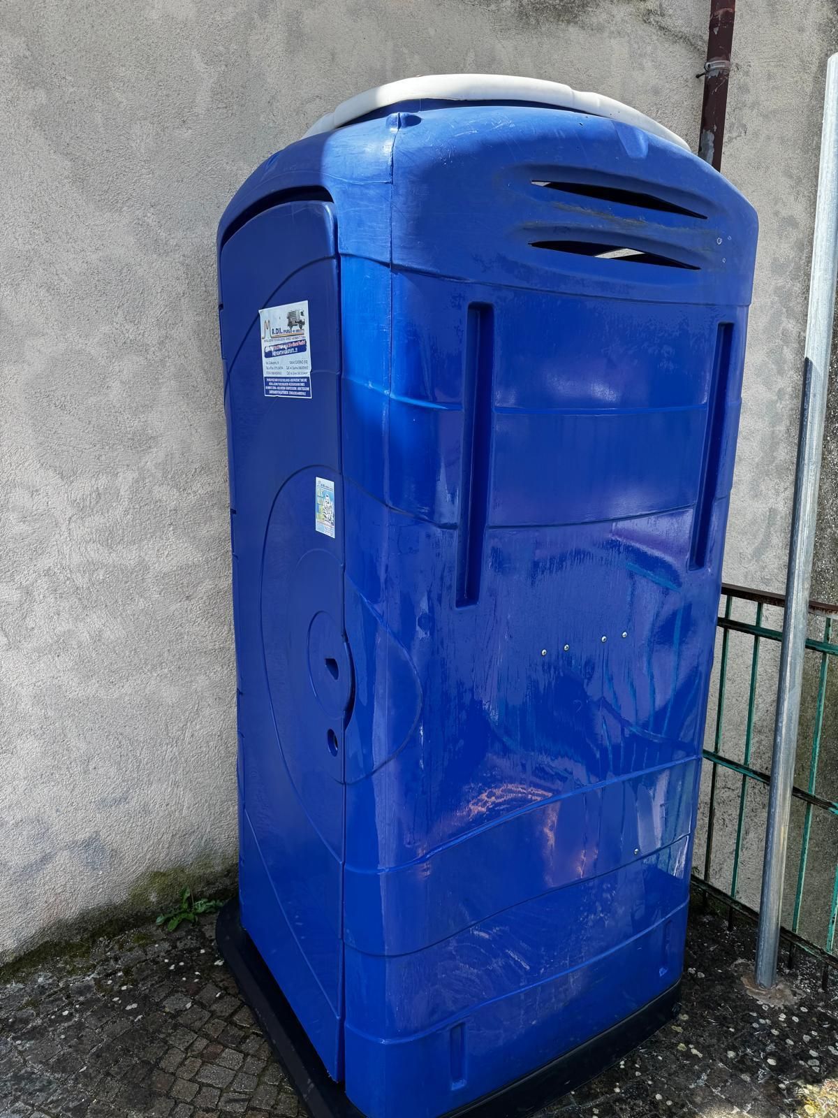 Contenitore blu per la raccolta differenziata accanto a un muro di cemento su una superficie esterna pavimentata.
