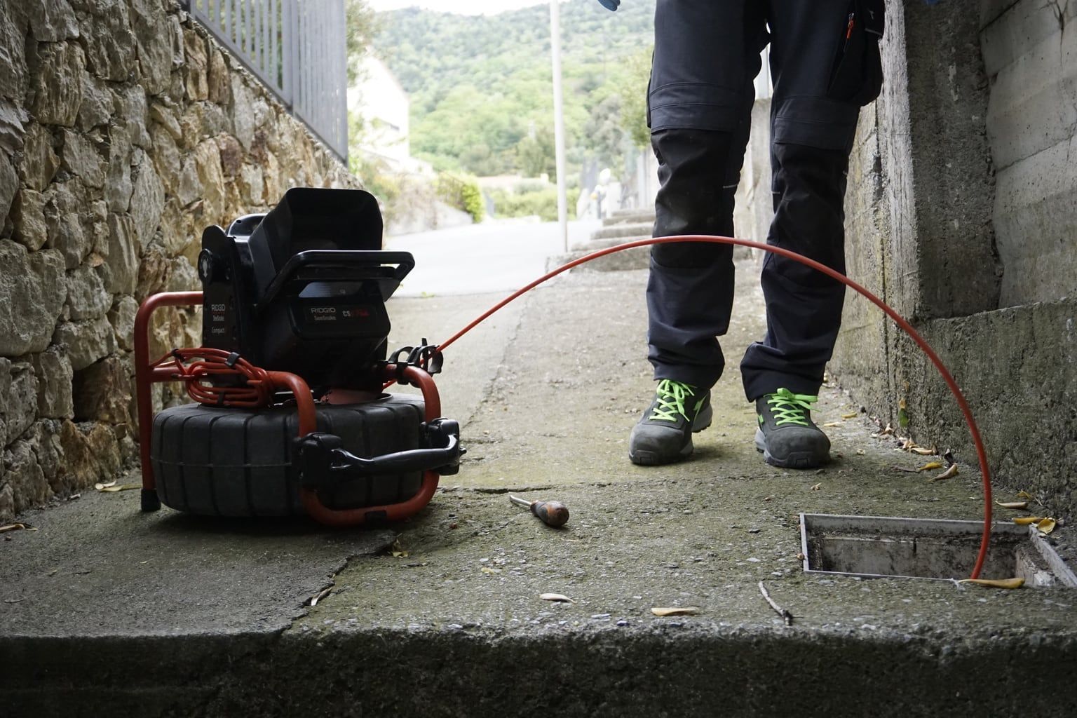 Un operaio è in piedi accanto a un avvolgitubo rosso e a un compressore in uno stretto vicolo di cemento.