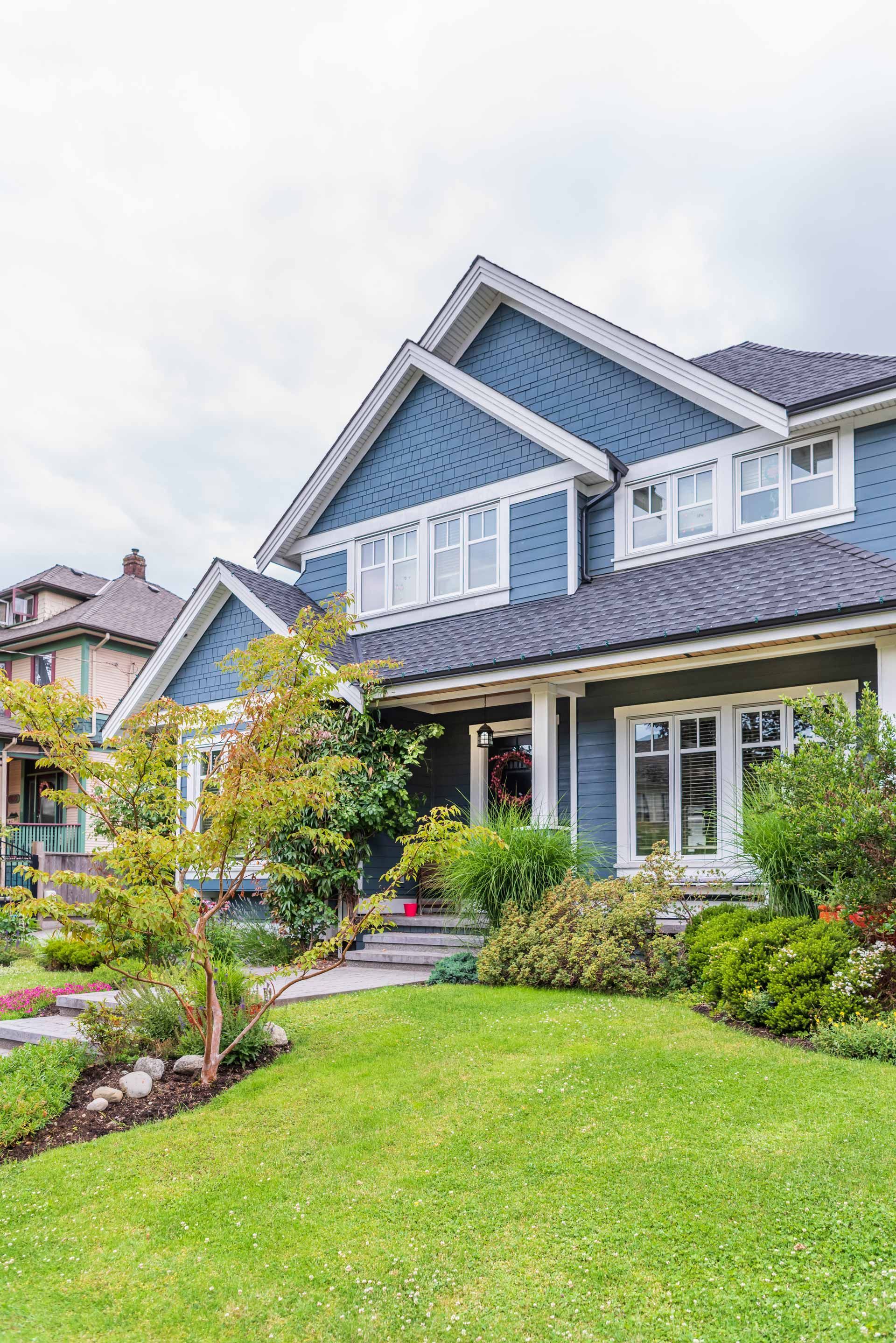 Two-story blue house with white trim, green lawn, and trees under a cloudy sky.