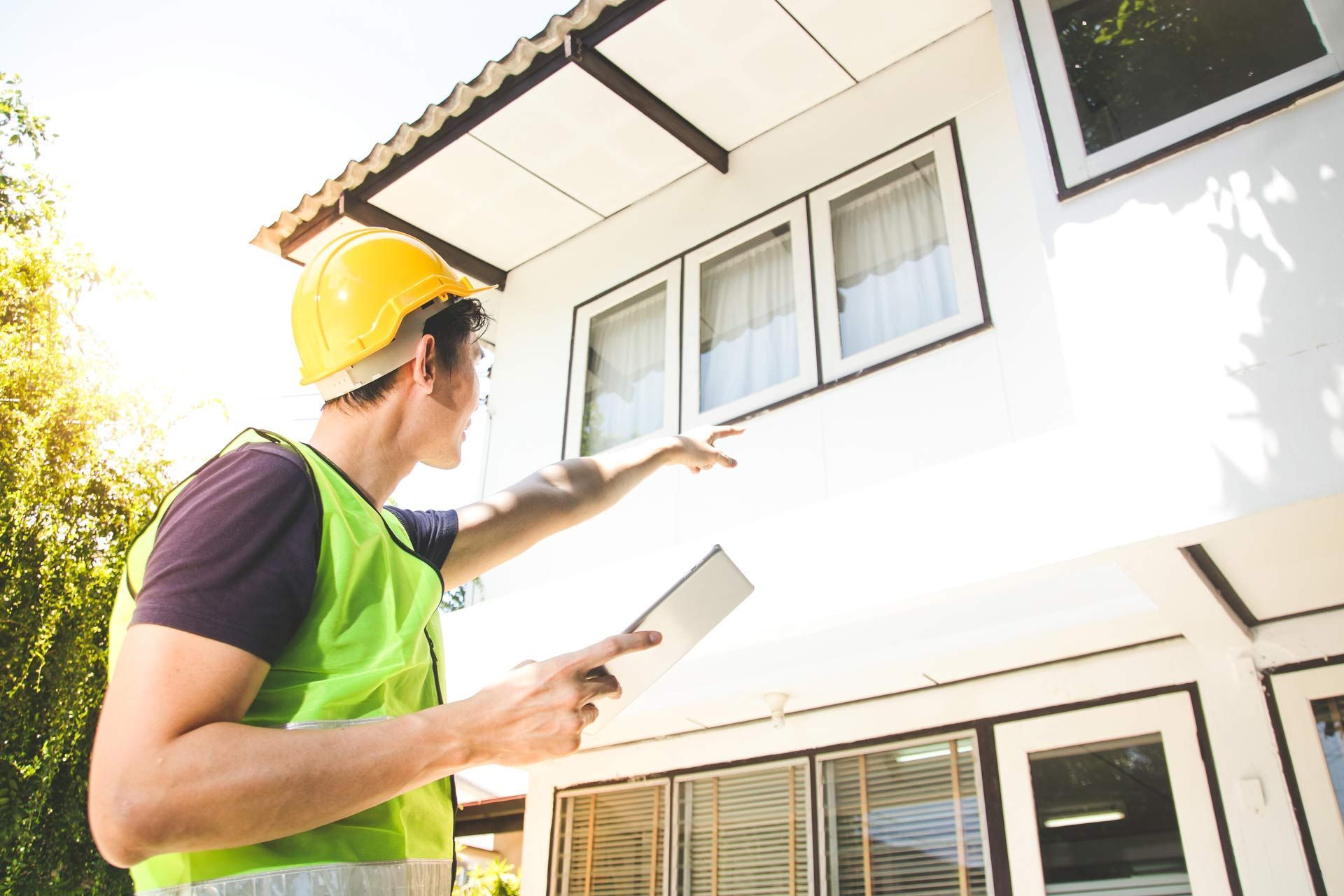 A construction worker is looking at a house while holding a tablet.