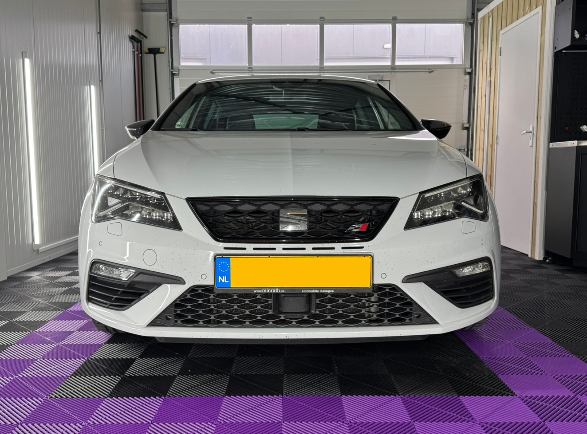 Een witte Seat Leon staat geparkeerd in een garage met een zwart-paars geblokte vloer.