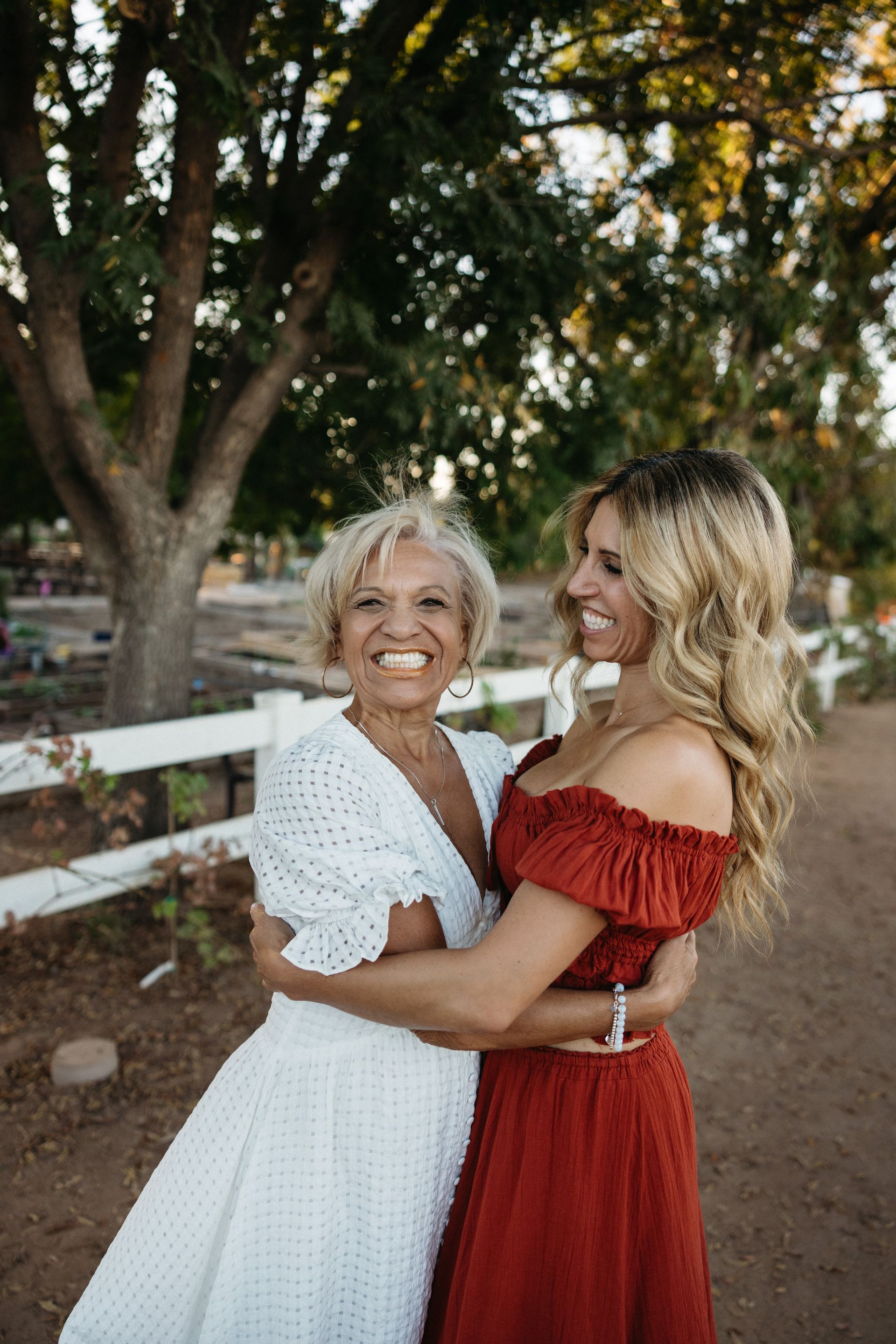 Woman in white dress hugs woman in red dress, smiling, outdoors near a white fence and tree.