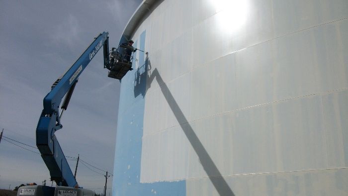Technician using a boom lift for precision tank painting on a large industrial storage unit.