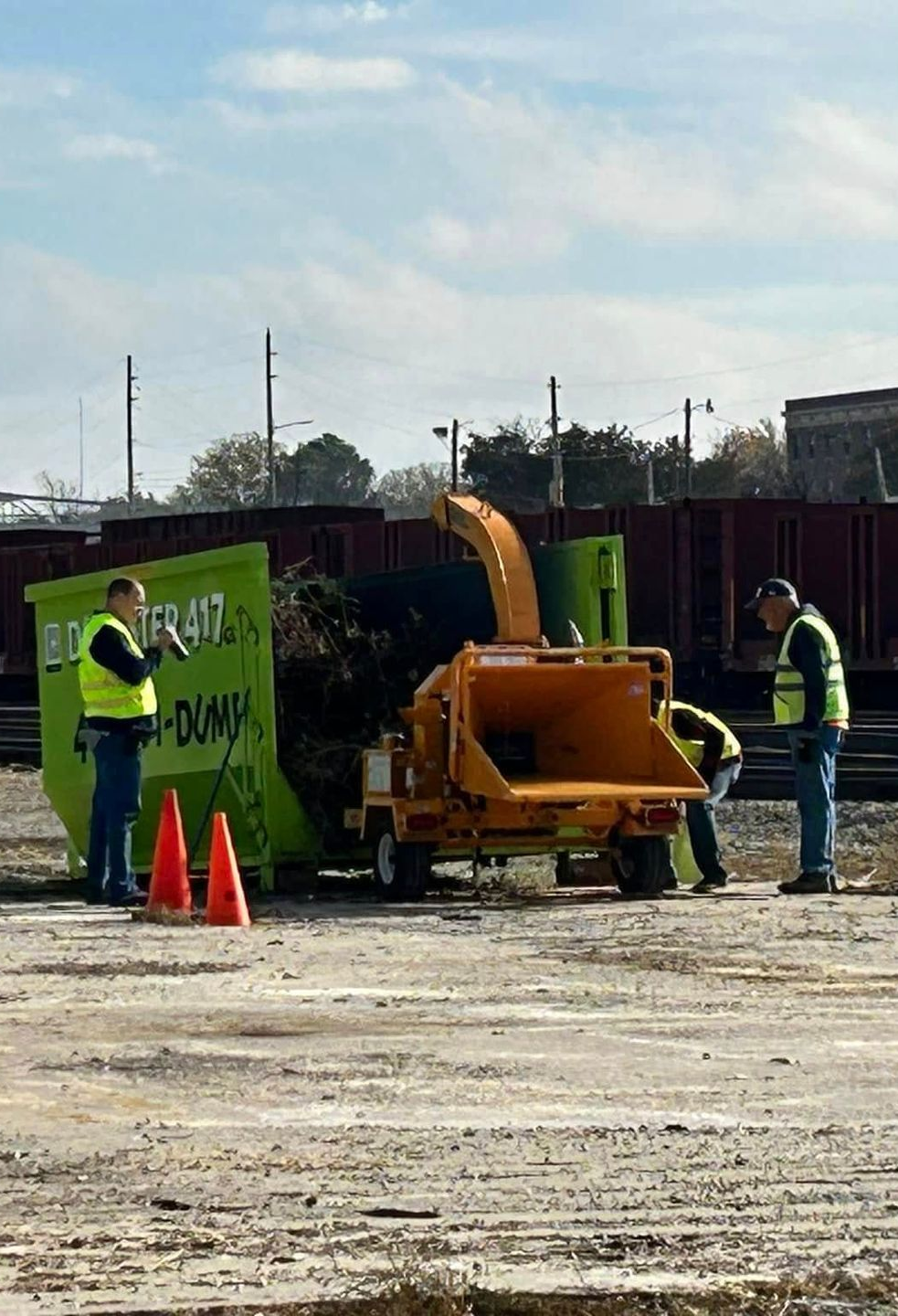 Construction workers around a yellow concrete pump at a muddy worksite with orange cones and green barriers