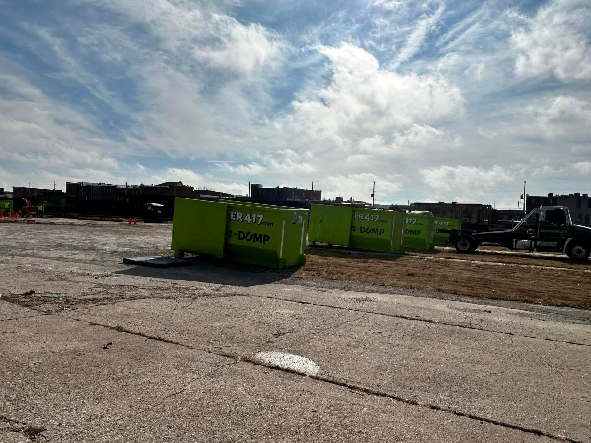 Bright green dumpsters in a dirt lot under a cloudy sky, with trucks parked nearby.