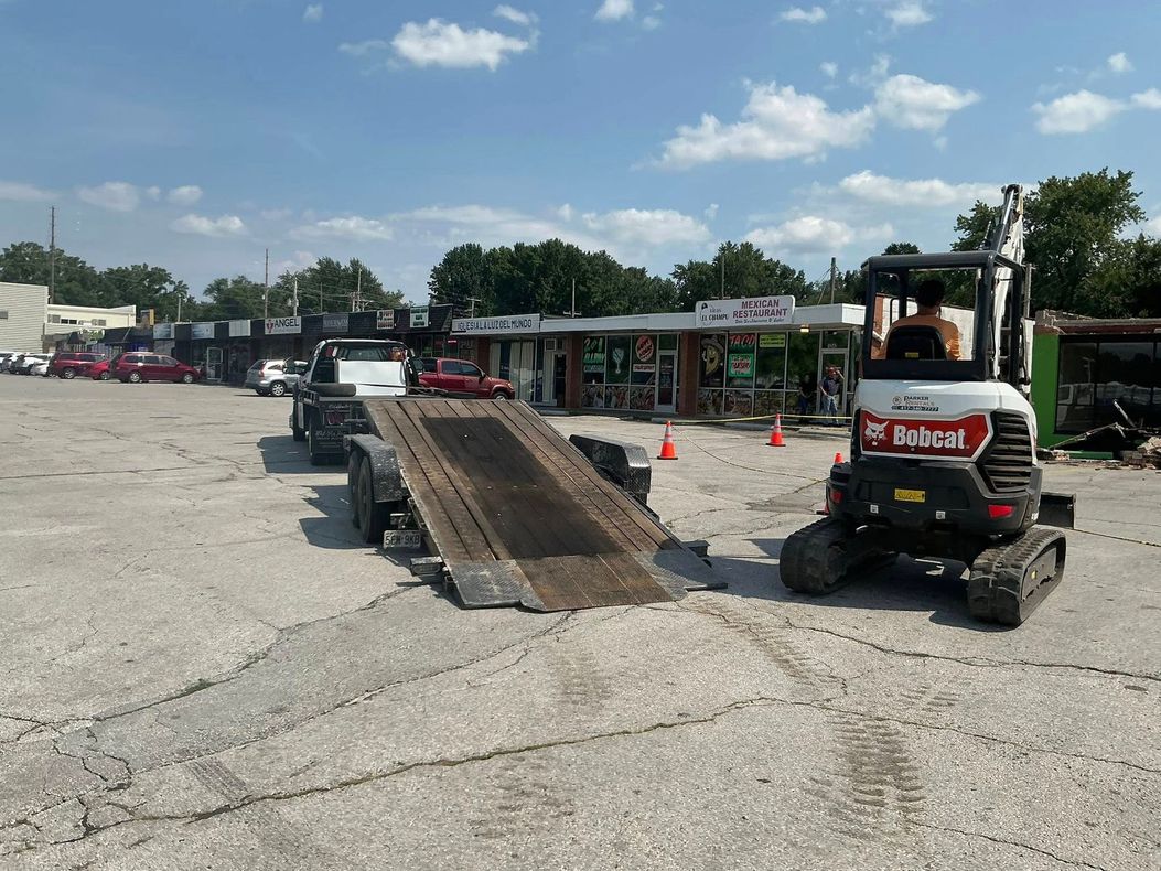 Bobcat skid steer on a dirt lot loading a metal ramp, with vehicles and buildings in the background.