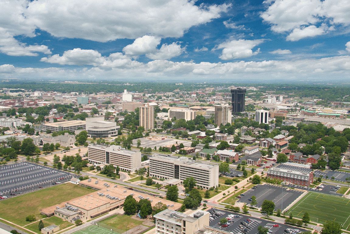 Aerial view of a city skyline with high-rise buildings, roads, trees, and a cloudy blue sky