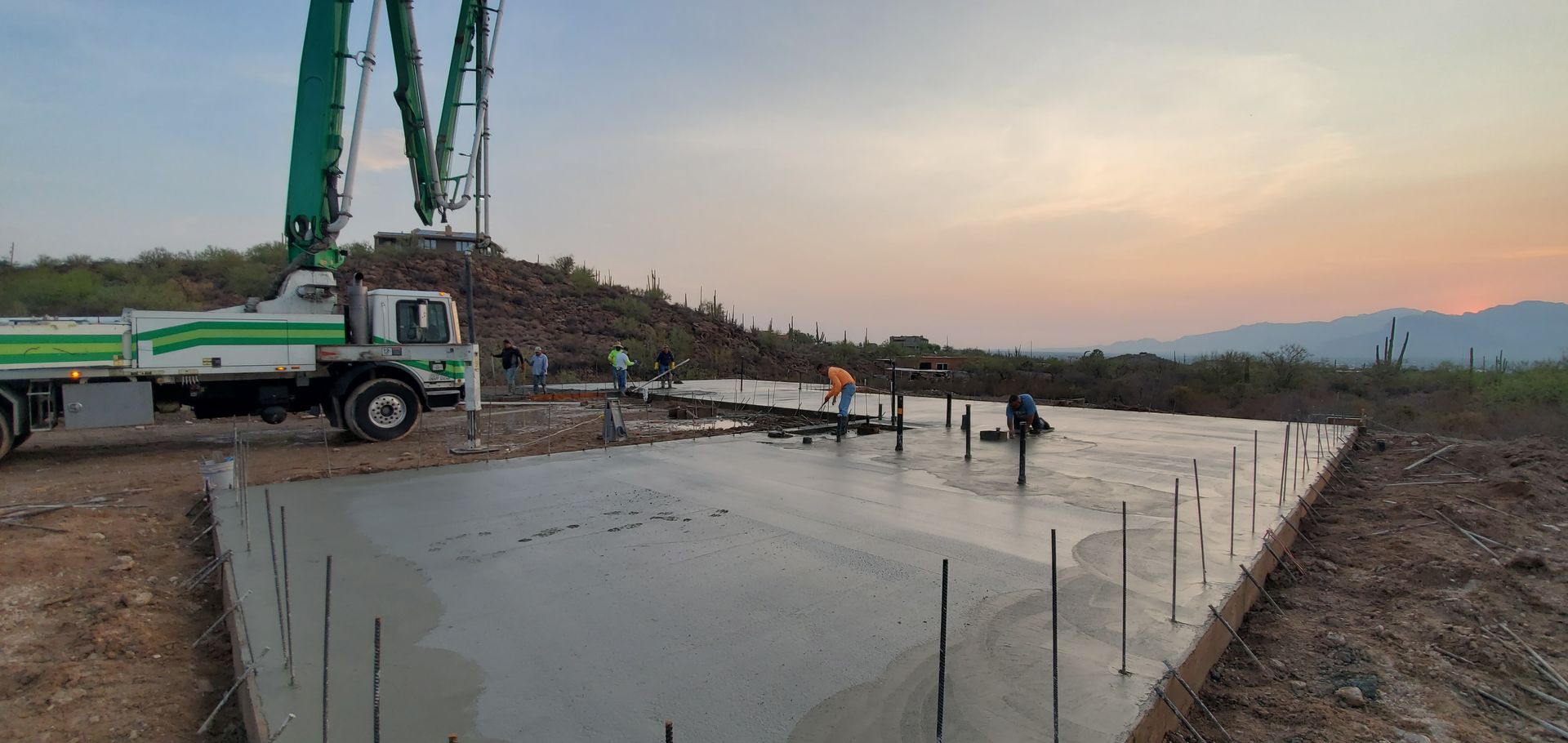 Concrete being poured for a foundation with a pump truck. Workers are present and the sky is dusky.
