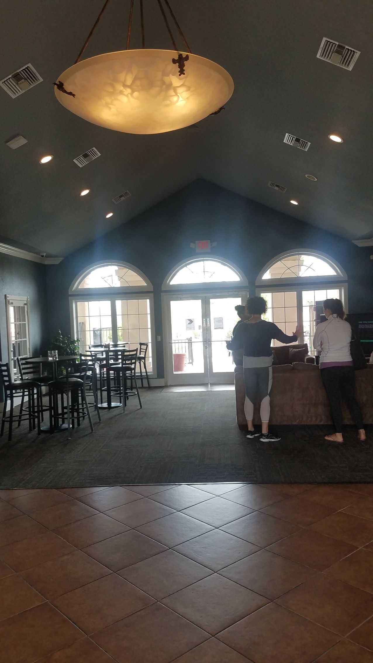 Interior with tables, arched windows, and people near a counter. Large light fixture hangs from the ceiling.