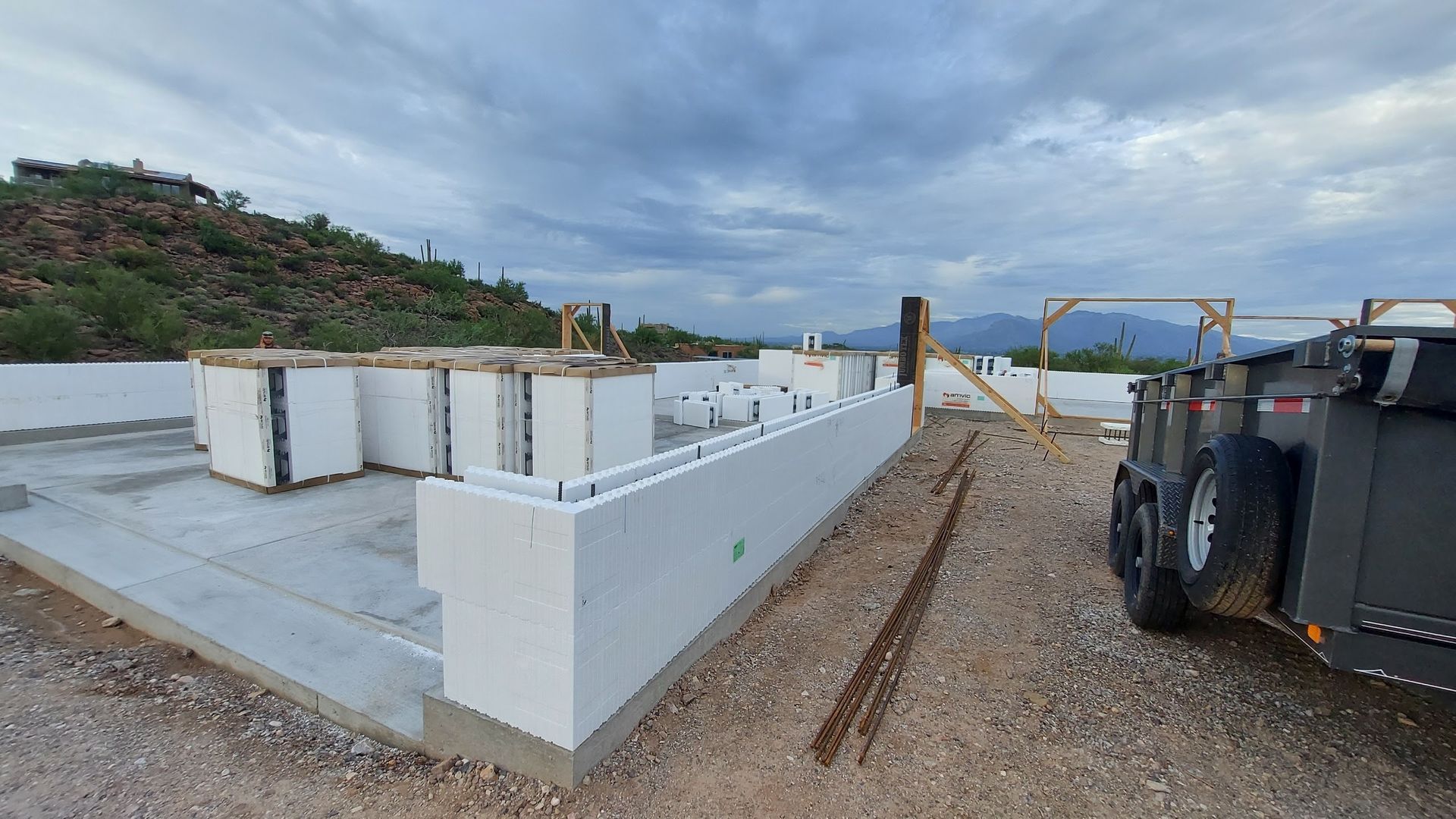 Construction site with white ICF blocks forming walls on a concrete foundation, mountains in the background, cloudy sky.