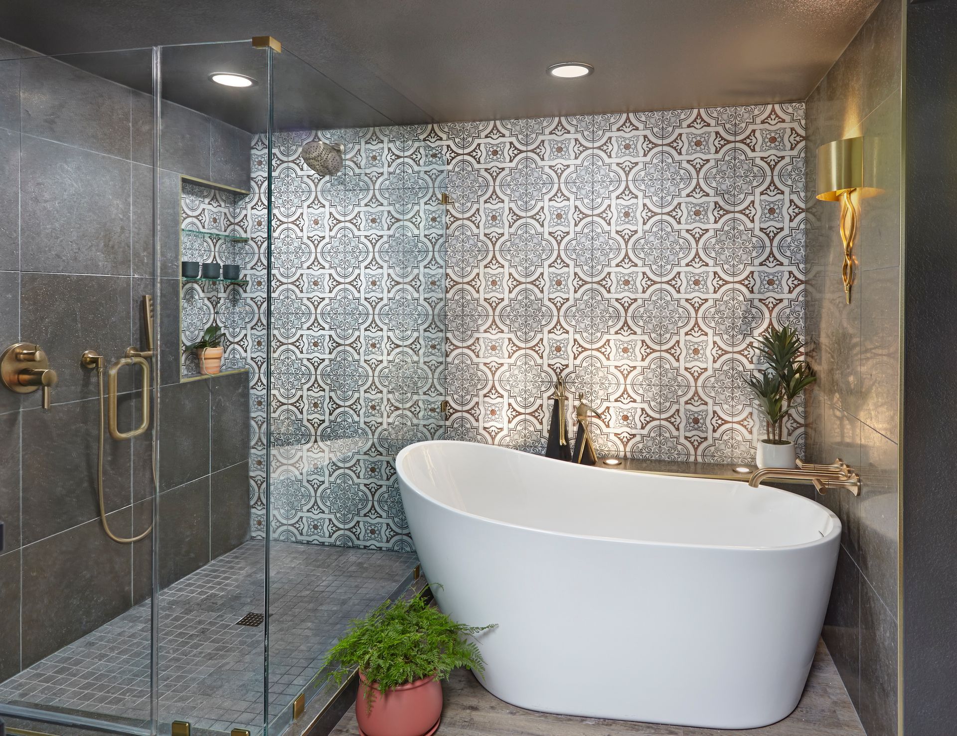 Modern bathroom with patterned tile accent wall, glass shower, freestanding white tub, and plants.