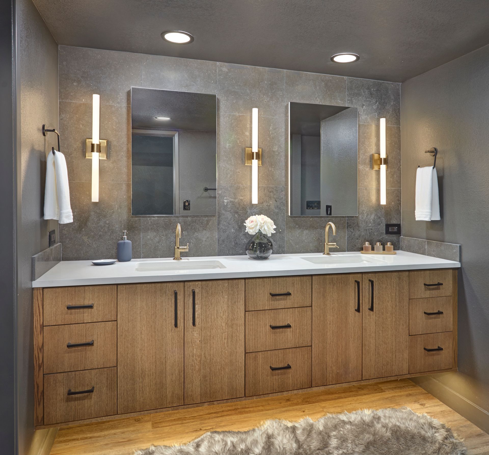 Bathroom vanity with two sinks, mirrors, and sconces; light wood cabinets, gray walls, and rug.