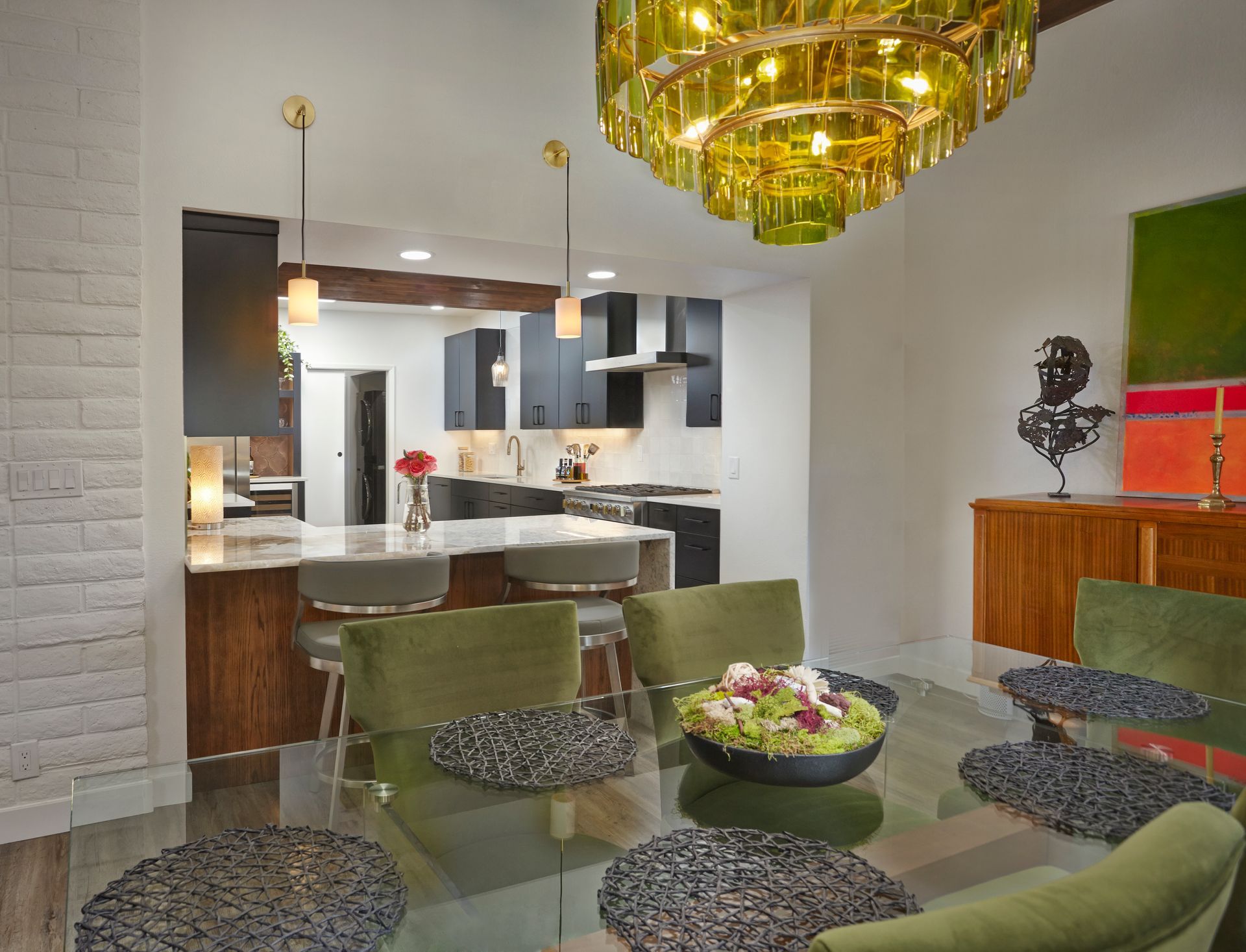 Dining room with open kitchen. Green chairs, glass table, golden chandelier, and brick accent wall.