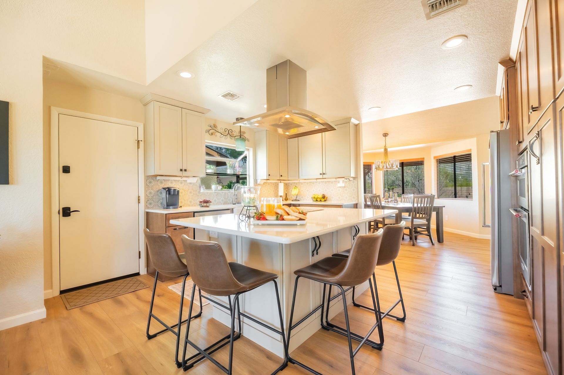 Modern kitchen with island, seating, stainless steel range hood, light cabinets, and wooden floors.