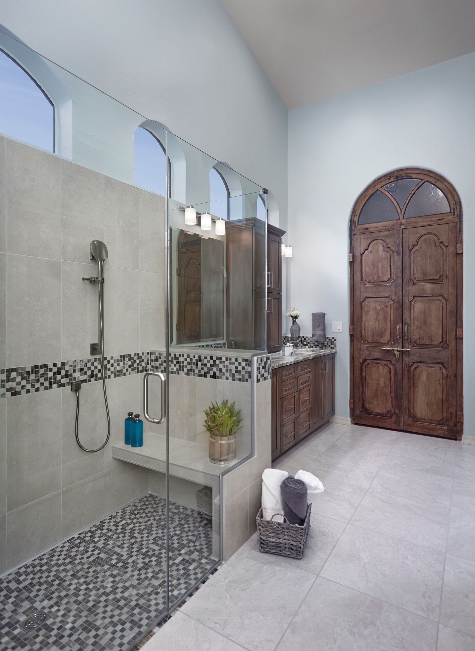Spacious bathroom with glass shower, wooden vanity, and large arched wooden door. Light blue walls and gray tile floor.