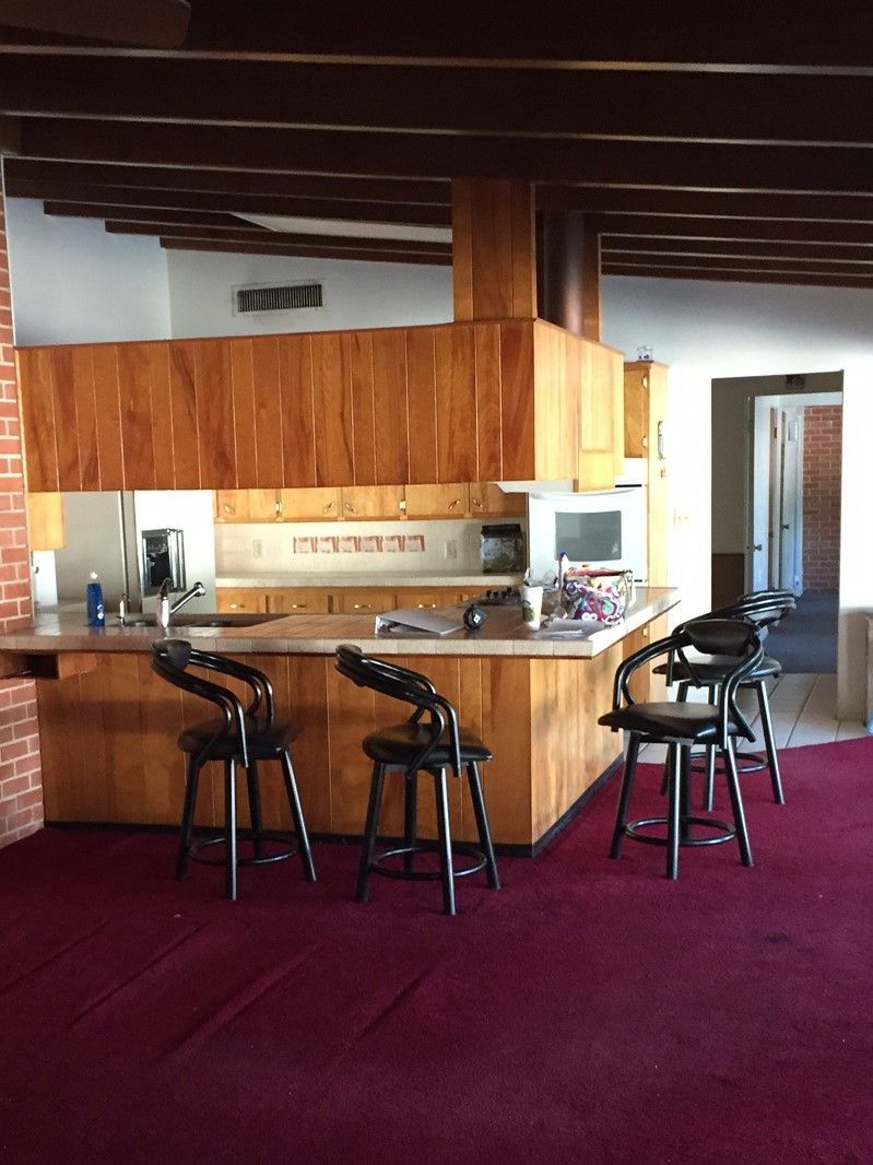 Kitchen with wooden cabinets, countertop, and bar stools; red carpet; brick wall; and recessed lighting.