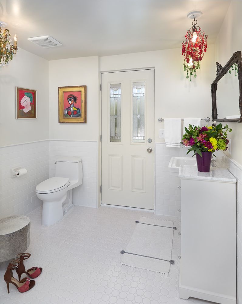 White bathroom with chandelier, toilet, art, door, and a vanity with flowers.