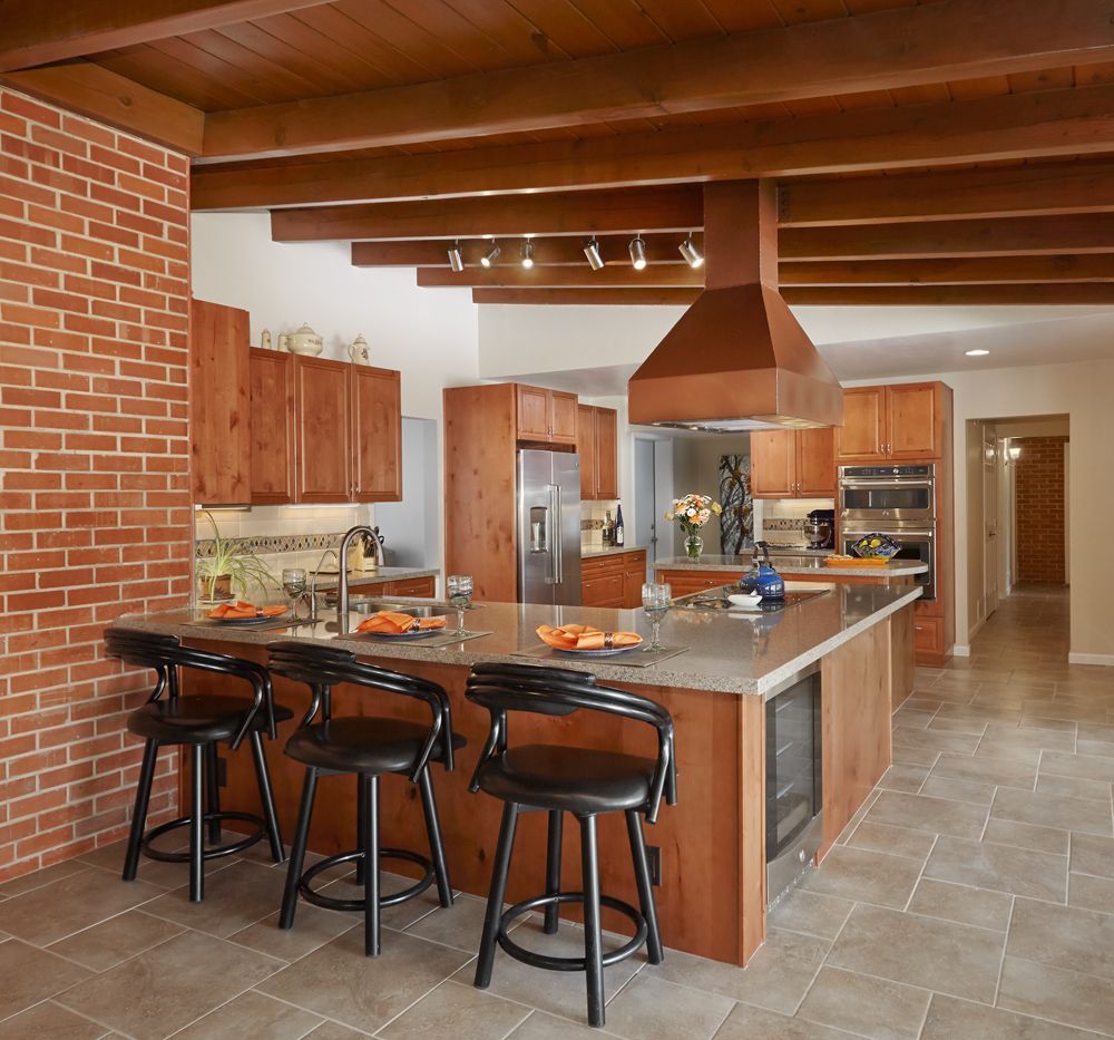 Kitchen with brick wall, wood cabinets, island with stools, and wooden beams.