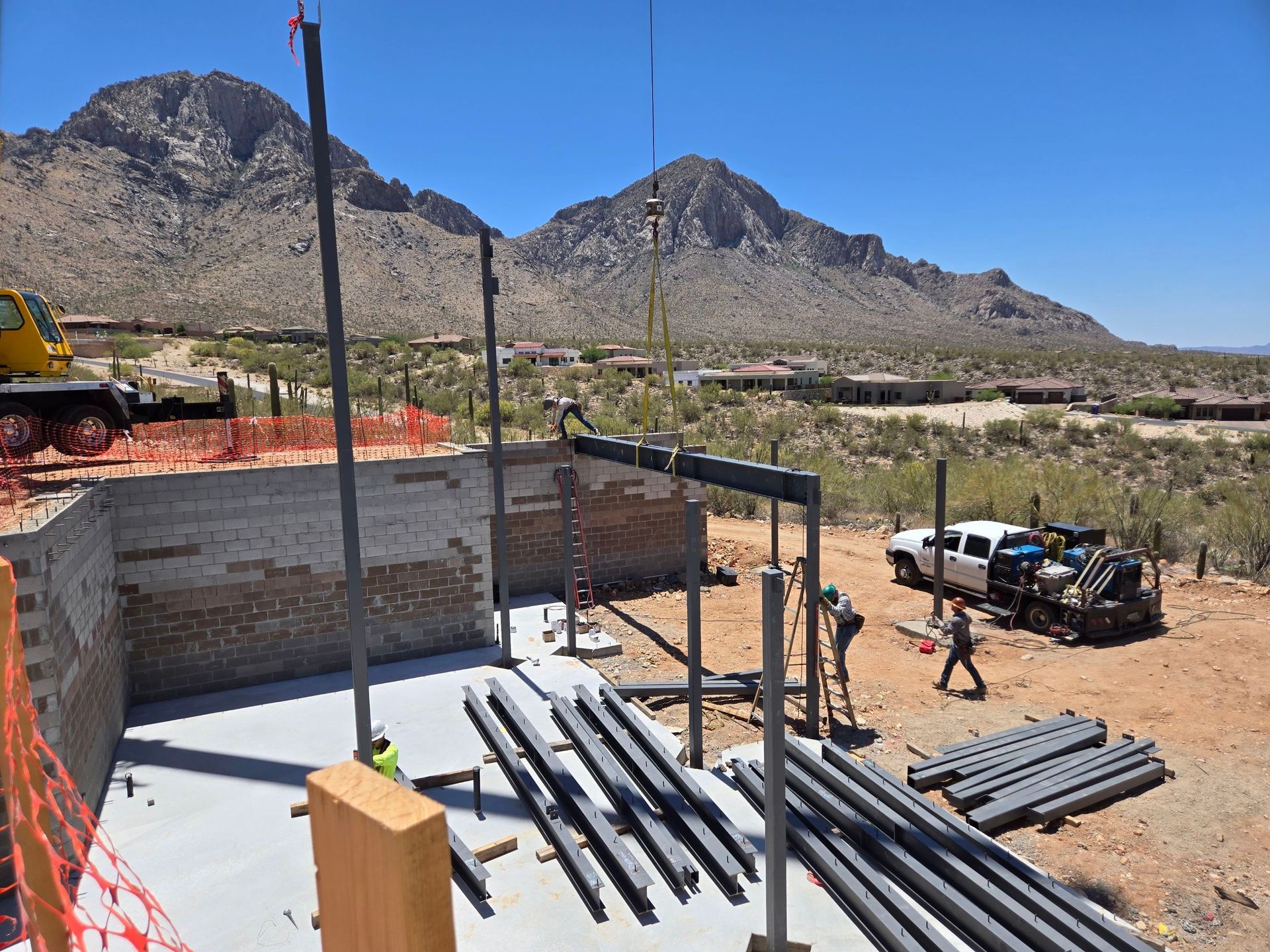 Construction site with steel beams being lifted into place by a crane, mountains in the background.