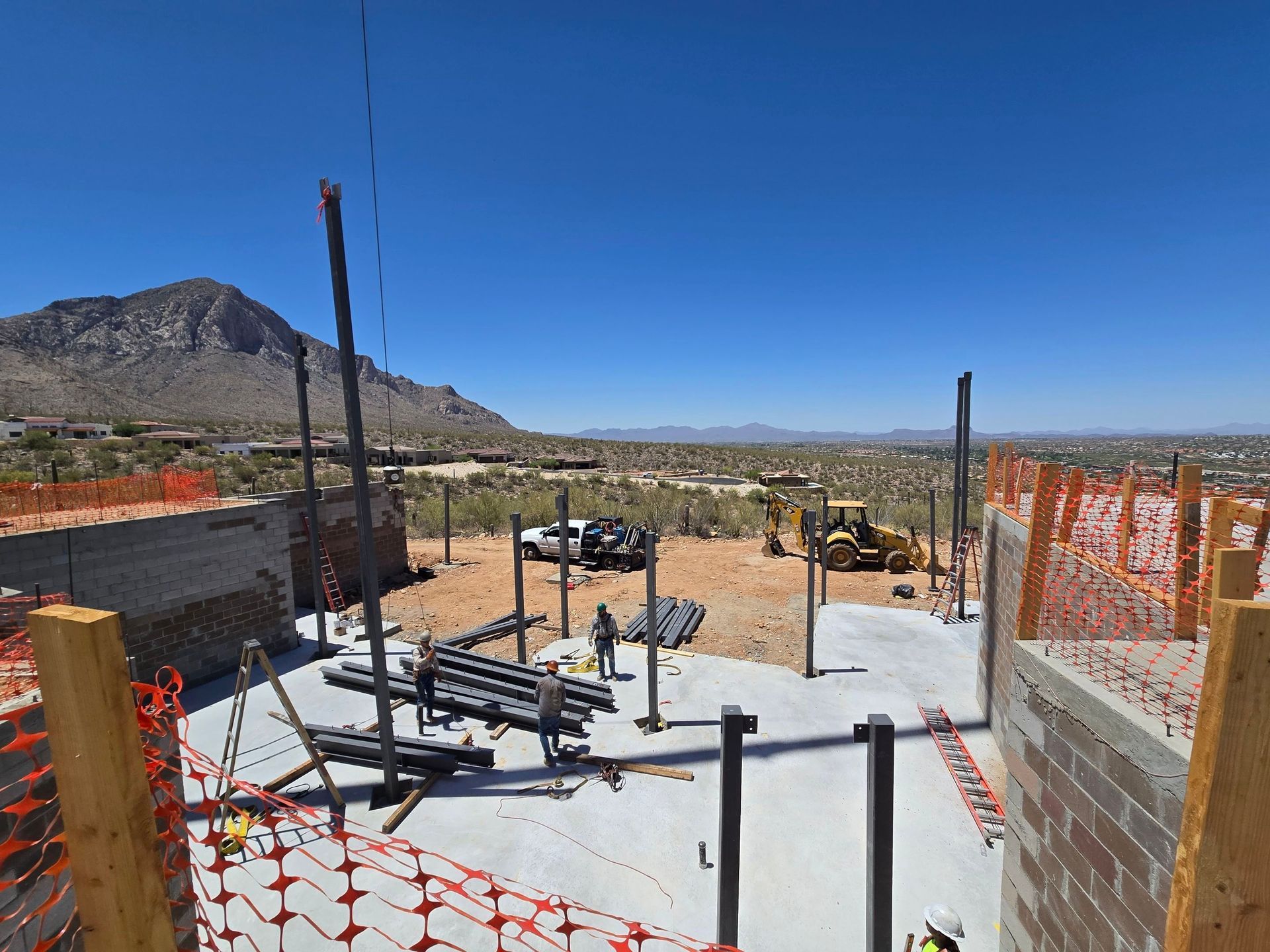 Construction site with concrete block walls, metal beams, safety fencing, and a mountain backdrop under a blue sky.