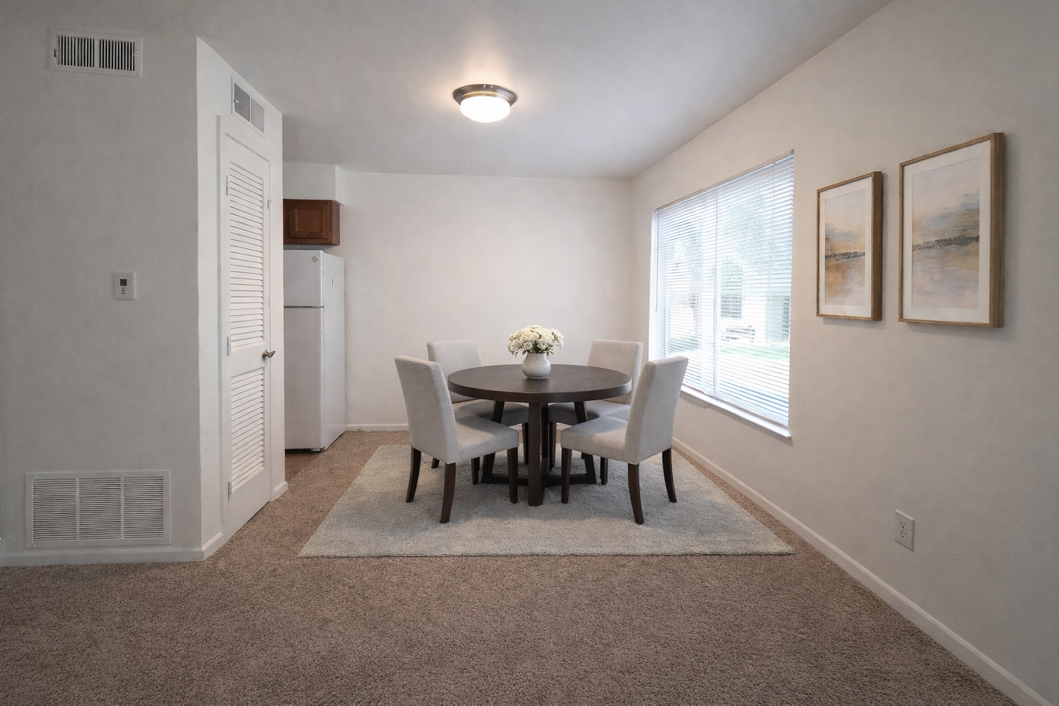 Dining area with round table, four gray chairs, window blinds, and framed wall art in a beige room