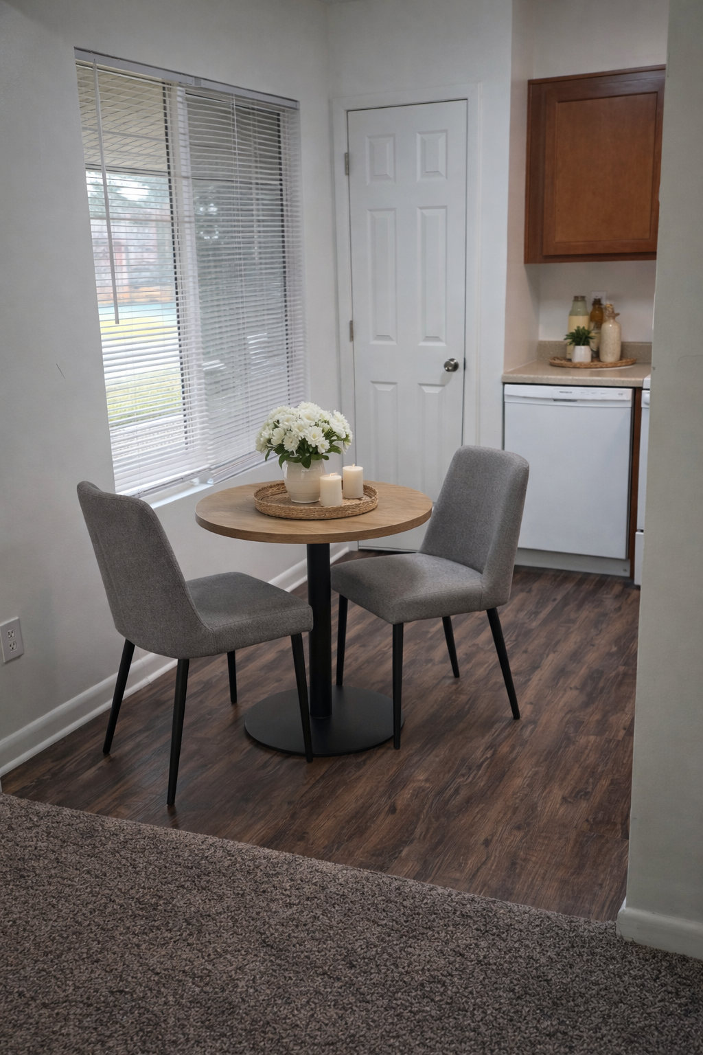 Small dining nook with round table, two gray chairs, white flowers, window, and kitchenette area