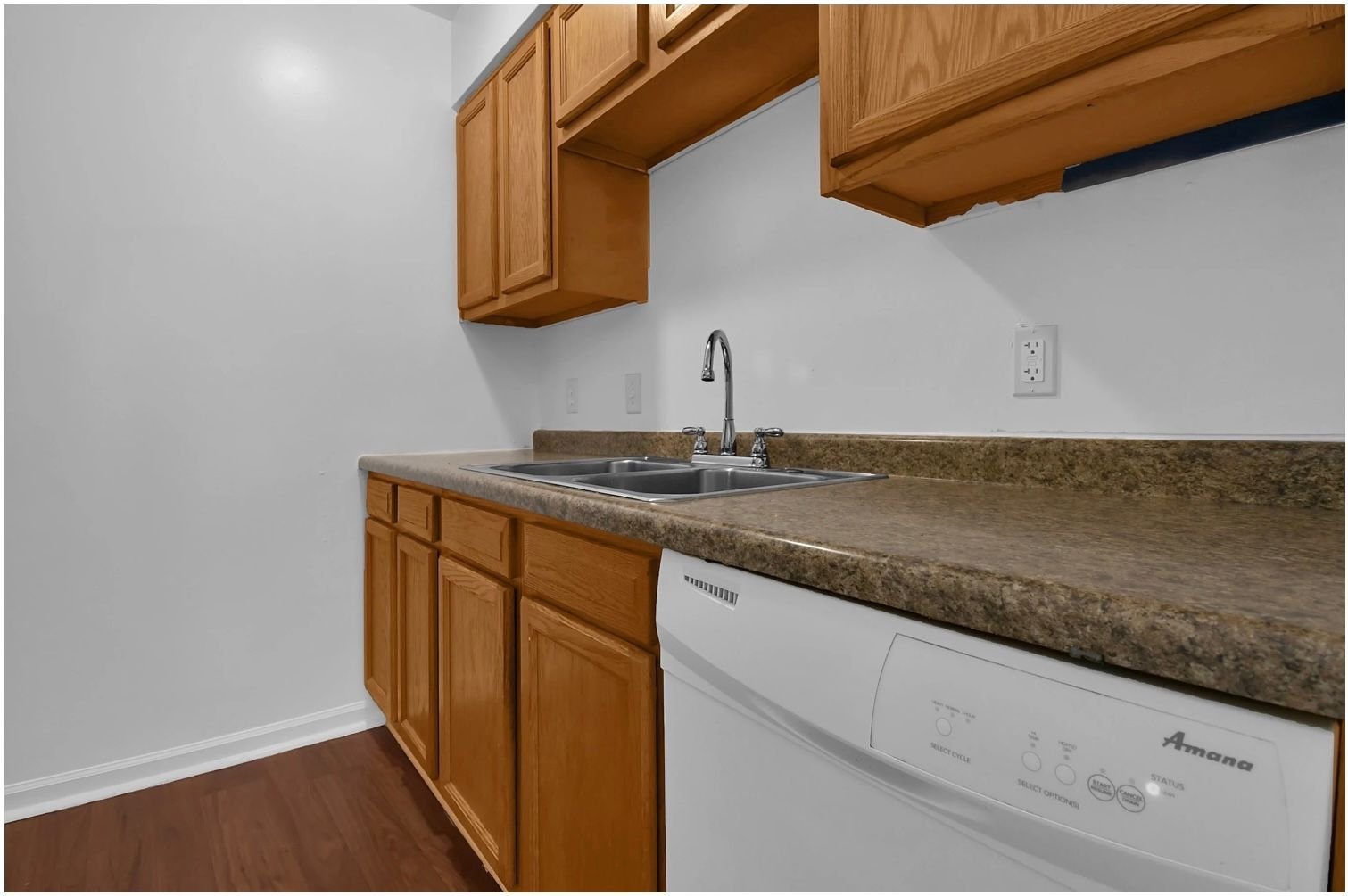 Small kitchen with wood cabinets, granite countertop, sink, and white dishwasher against gray walls.