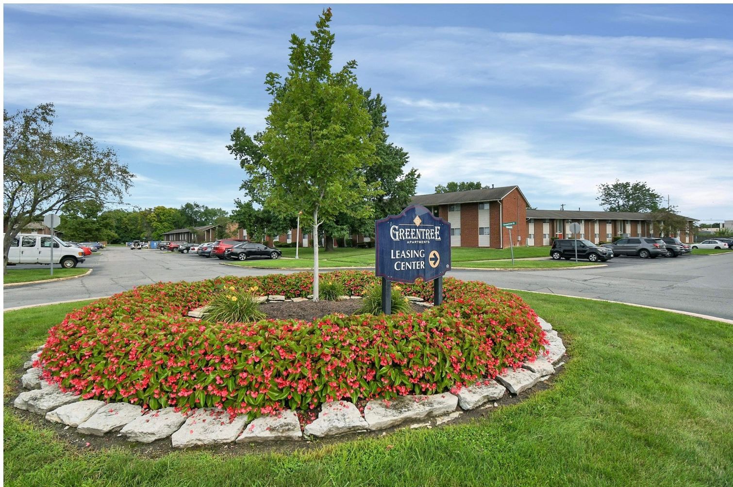 Landscaped apartment entrance with flower bed, sign, trees, and brick buildings under a cloudy sky