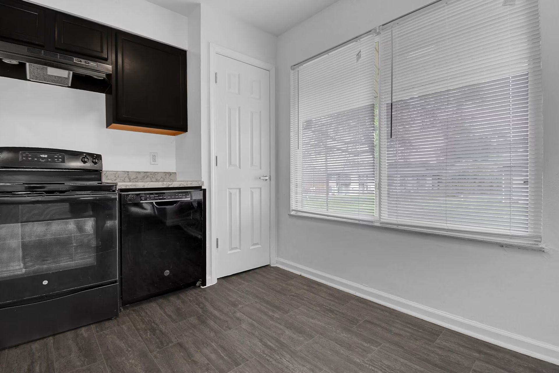 Empty kitchen with black appliances, dark wood floor, and large window with closed blinds.