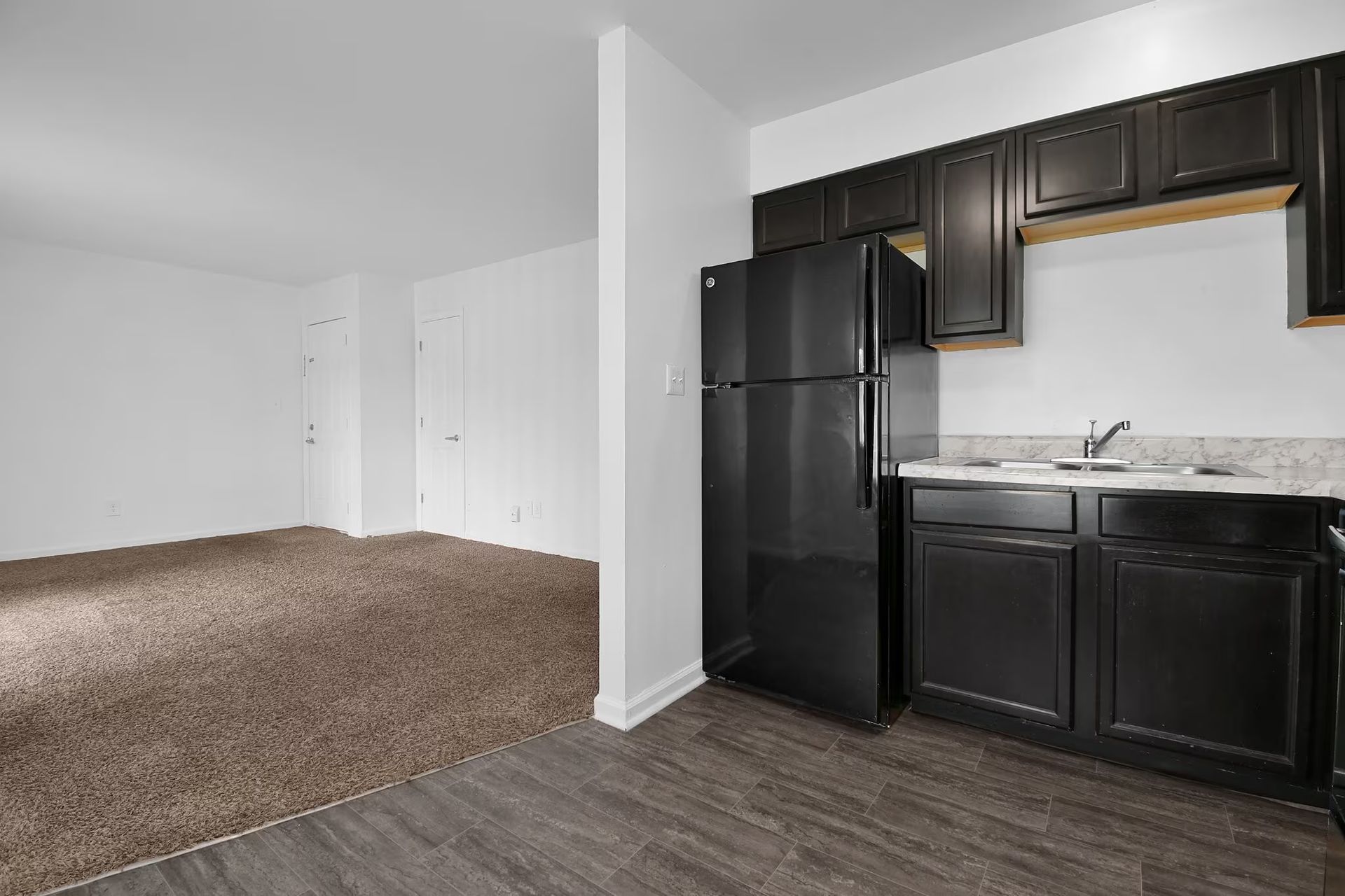 Empty apartment living area with carpet and black kitchen cabinets, refrigerator, and dark wood flooring