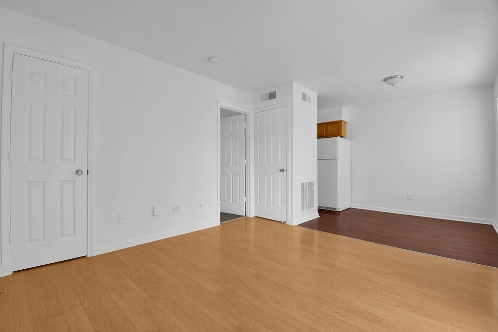 Empty white room with wood flooring, white doors, and a small brown carpeted area in the back corner