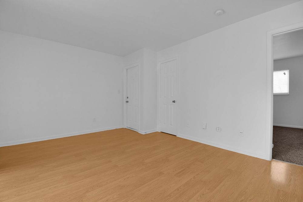Empty white room with tan flooring, two closed doors, and an open doorway to a bathroom