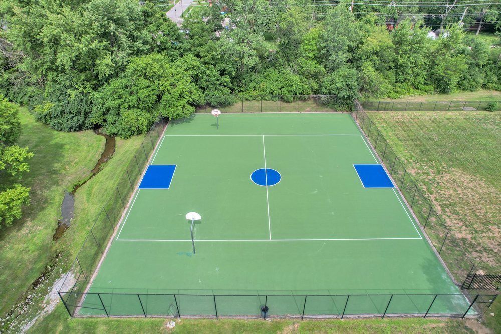 Empty green sports court with blue center markings, surrounded by trees and a fence