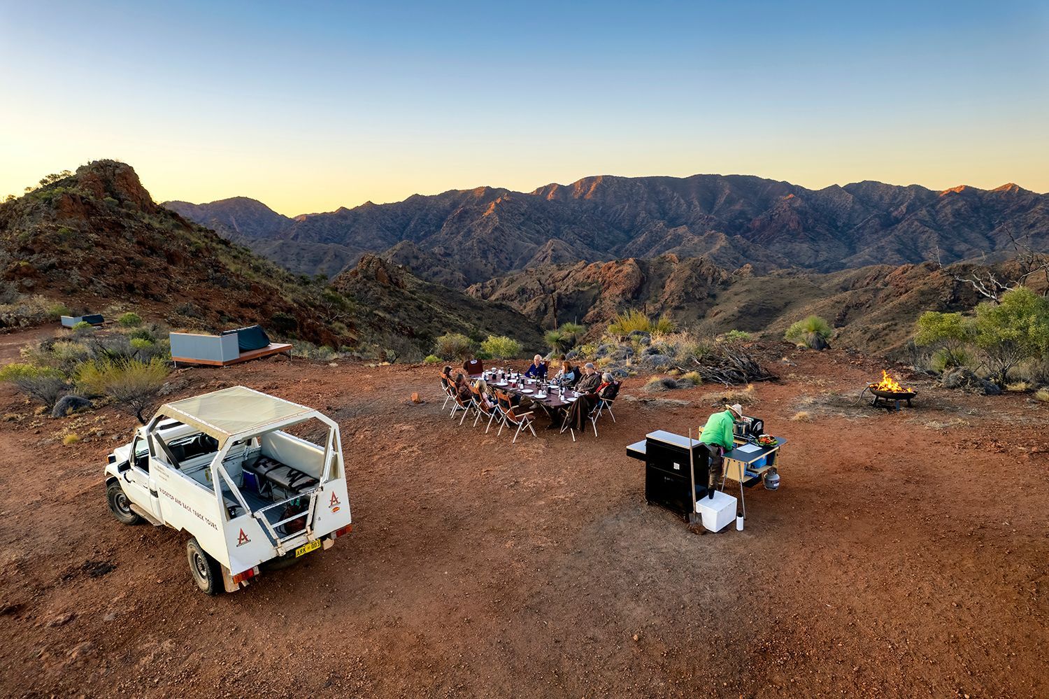 Group of people enjoying outdoor dining at sunset amidst the rugged mountain ranges of Arkaroola, highlighting the unique Ridgetop Sleepout experience in the Australian outback. Experience this on the Arkaroola Photography Tour.