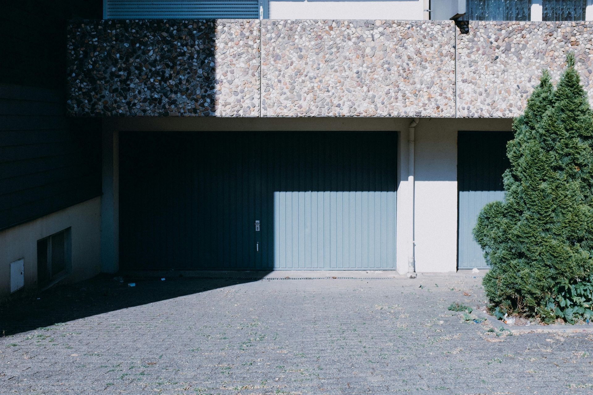 Garage with blue door under a concrete balcony, gravel driveway, and a small evergreen tree.