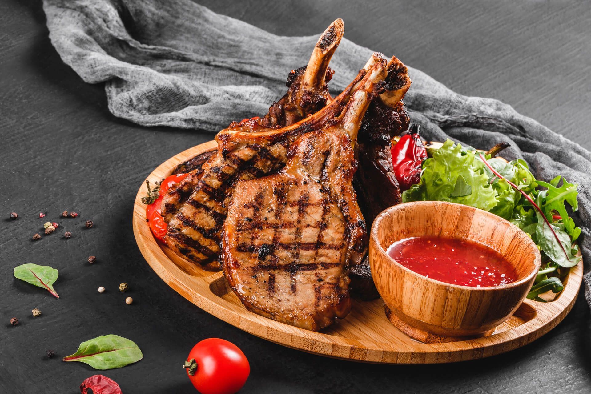 A wooden plate topped with grilled lamb chops and a bowl of sauce