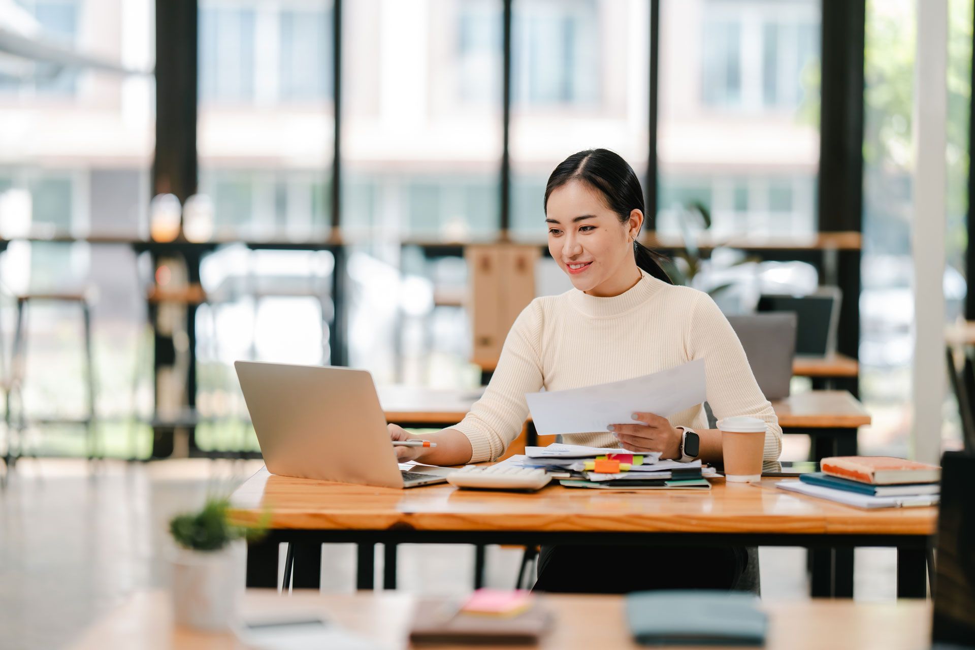 Woman smiling at a laptop, holding papers, at a desk in a bright office.