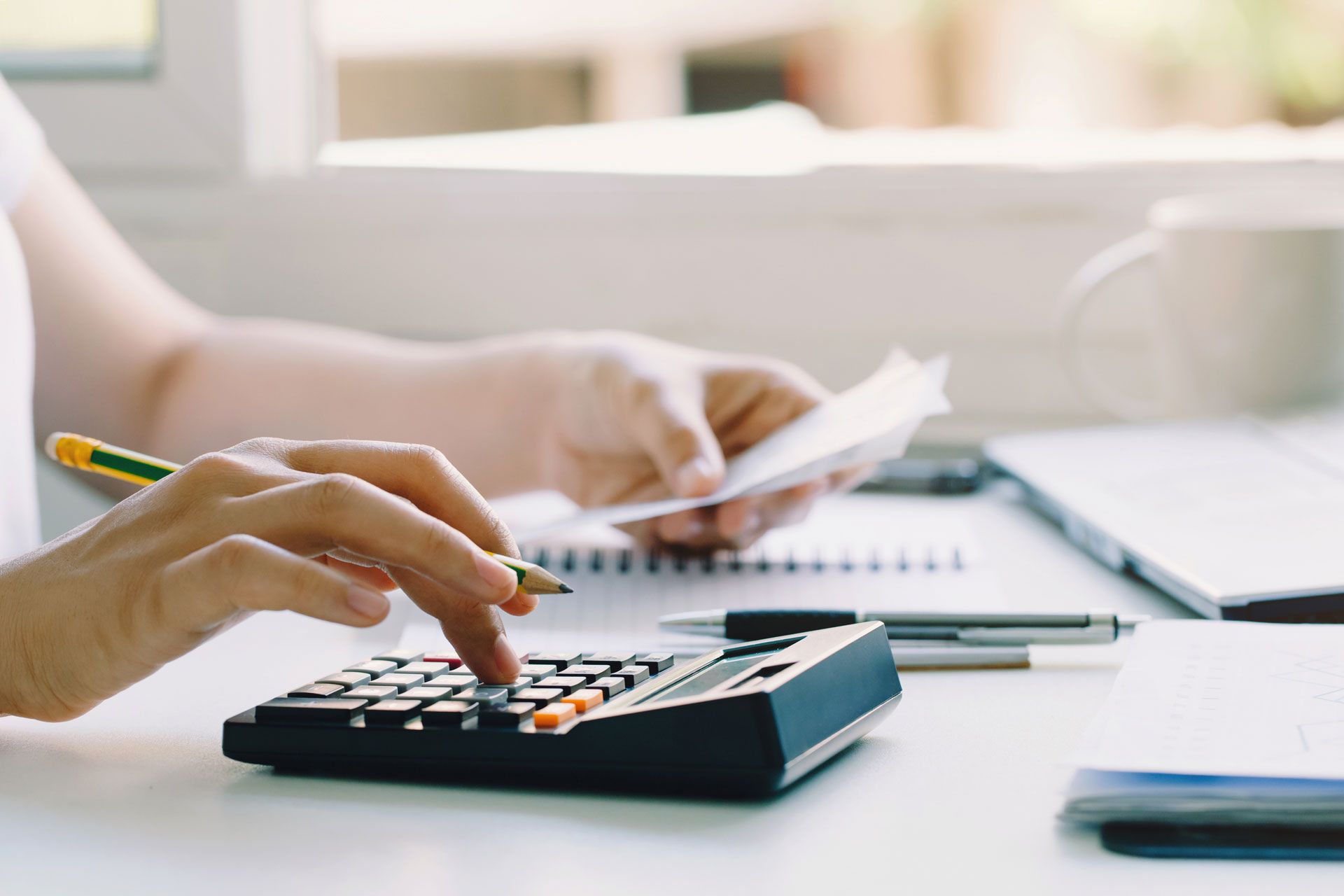 Person calculating expenses with a calculator and receipts on a desk near a window.