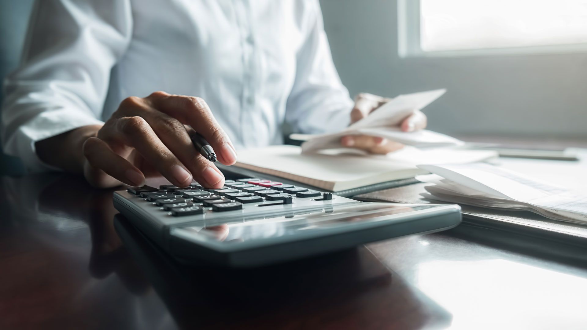 Person in white shirt using a calculator, holding receipts, and writing in a notebook at a desk.
