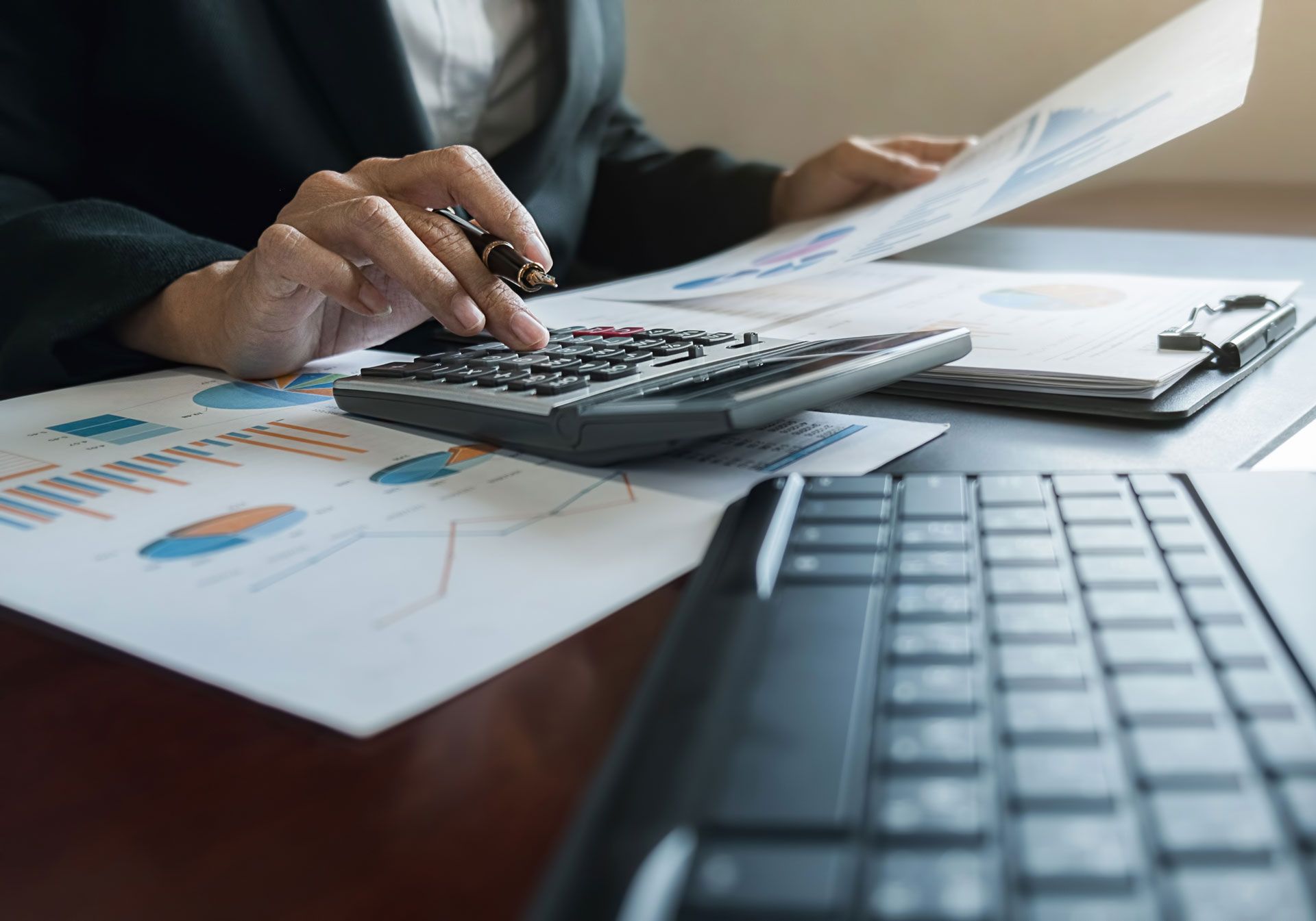 Person in suit using a calculator, reviewing financial charts and a laptop on a desk.