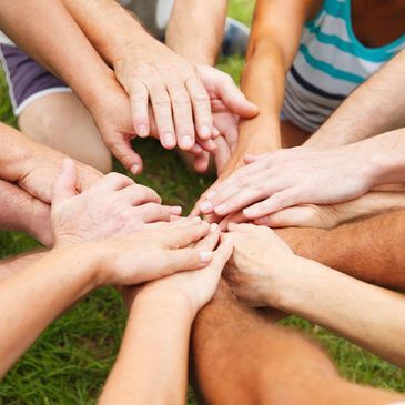 A group of people placing their hands on top of one another in a circle over a grassy background.