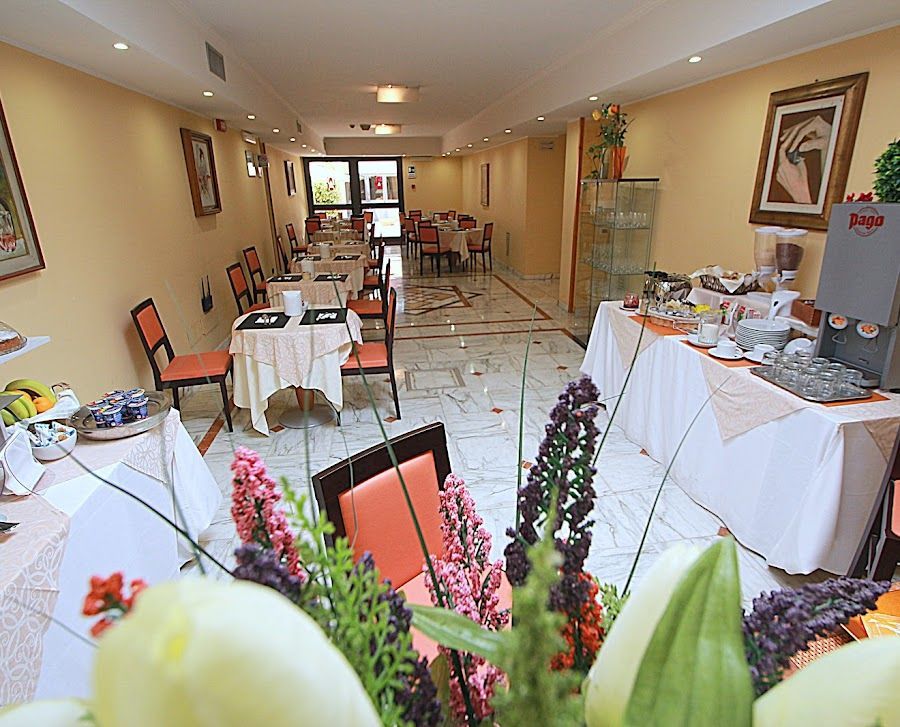 Sala colazioni dell'hotel con tavoli, sedie e buffet. Pareti gialle, tovaglie bianche e composizioni floreali.